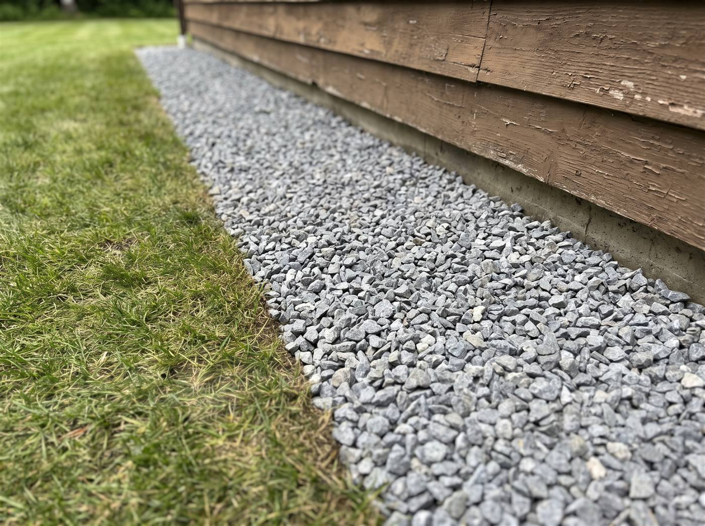 A clean strip of grey gravel running along the foundation of a house, with a sharp edge where it meets the grass beyond